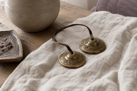 Deux cymbales tingsha en laiton doré reliées par une lanière de cuir, posées sur du lin.