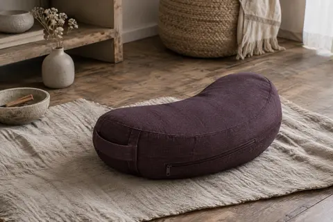 Coussin de méditation en croissant aubergine, avec housse texturée et poignée, posé sur un tapis de lin.