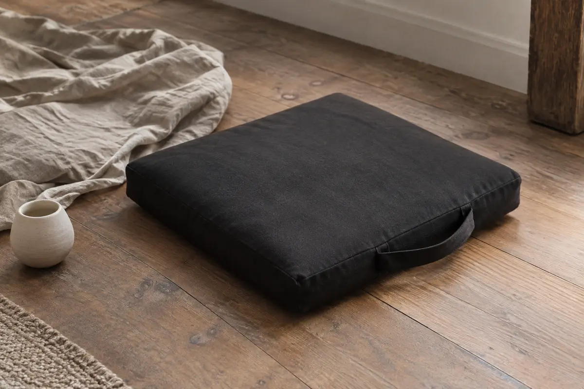 Zabuton carré noir en coton posé sur un parquet, avec sa sangle de transport assortie visible