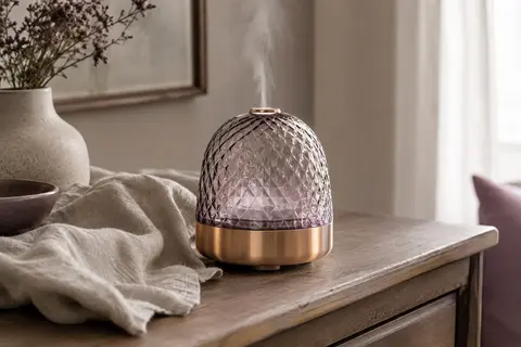Diffuseur d’arômes au couvercle en verre quadrillé et à la base or rose, posé sur une console en bois.