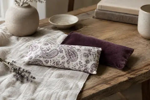 Coussin pour les yeux blanc avec housse violette posé sur du lin froissé près d’une branche de lavande.