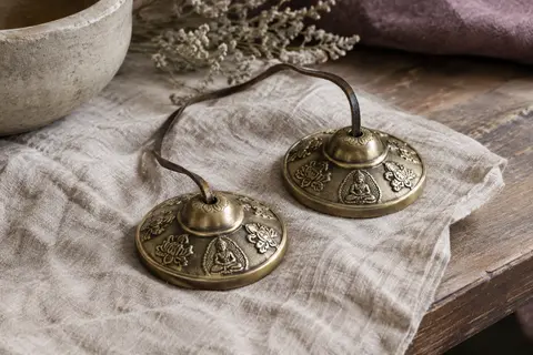 Tingsha tibétaine en bronze aux motifs bouddhistes, reliée par une corde en cuir et posée sur du lin.