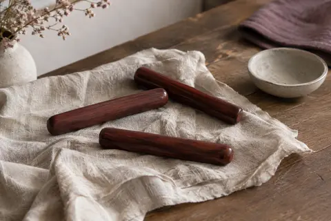 Trois bâtons de massage en bois rouge foncé posés sur du lin froissé près d’une coupelle en céramique.