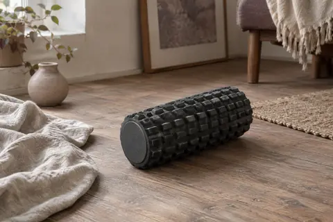 Rouleau de massage noir TriggerPoint Grid 1.0 à relief texturé posé sur un sol en bois près d’un linge de lin.