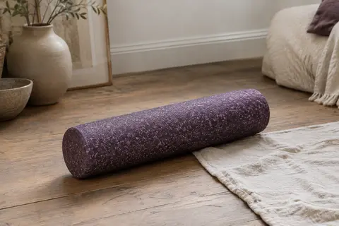 Rouleau de massage cylindrique pourpre moucheté posé sur un tapis en lin dans un intérieur calme.
